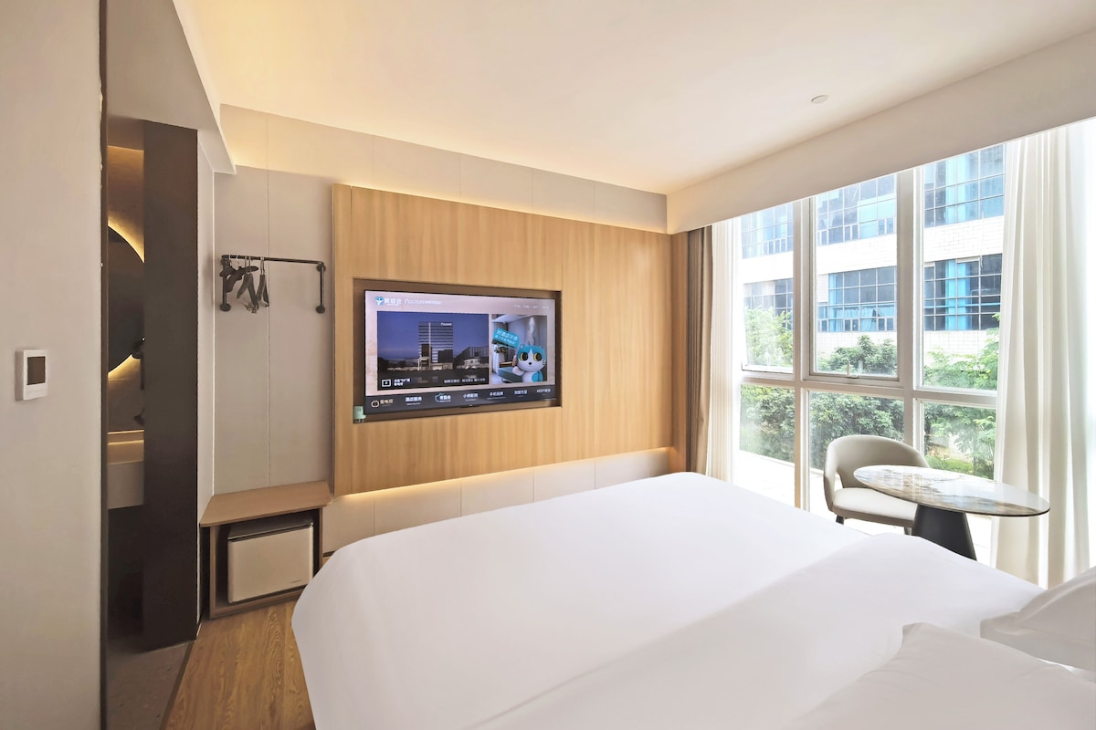 Hotelkamer met een bed, een aan de muur bevestigde televisie, een klein tafeltje en een raam met uitzicht op gebouwen en groen.