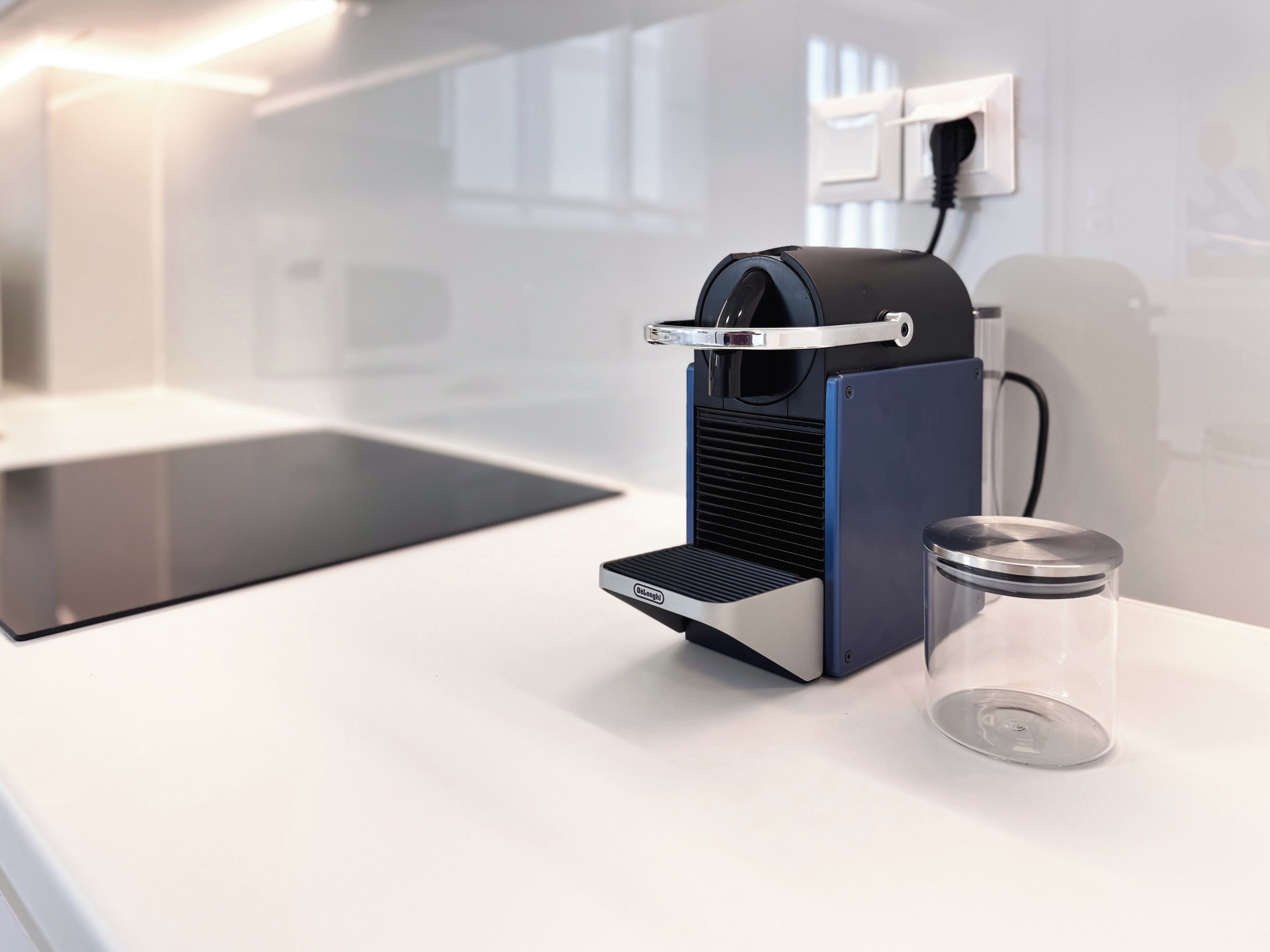 Una máquina de café con un recipiente de vidrio transparente a su lado, un enchufe y una encimera de cocina moderna.