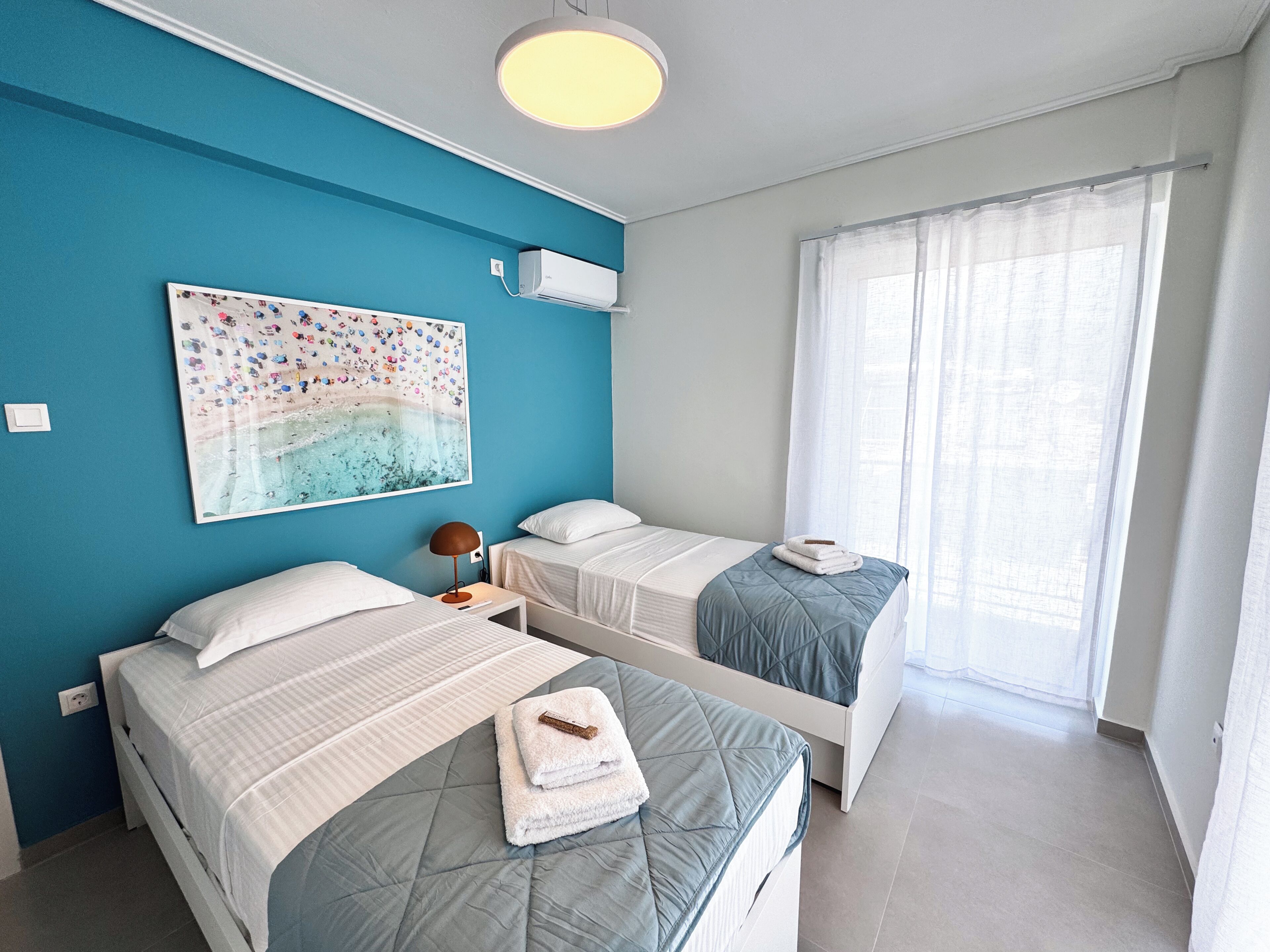 Een kamer met twee bedden, een plafondlamp, een wandairconditioner, een raam met dunne gordijnen en een schilderij met een strandthema.