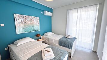 Een kamer met twee bedden, een plafondlamp, een wandairconditioner, een raam met dunne gordijnen en een schilderij met een strandthema.