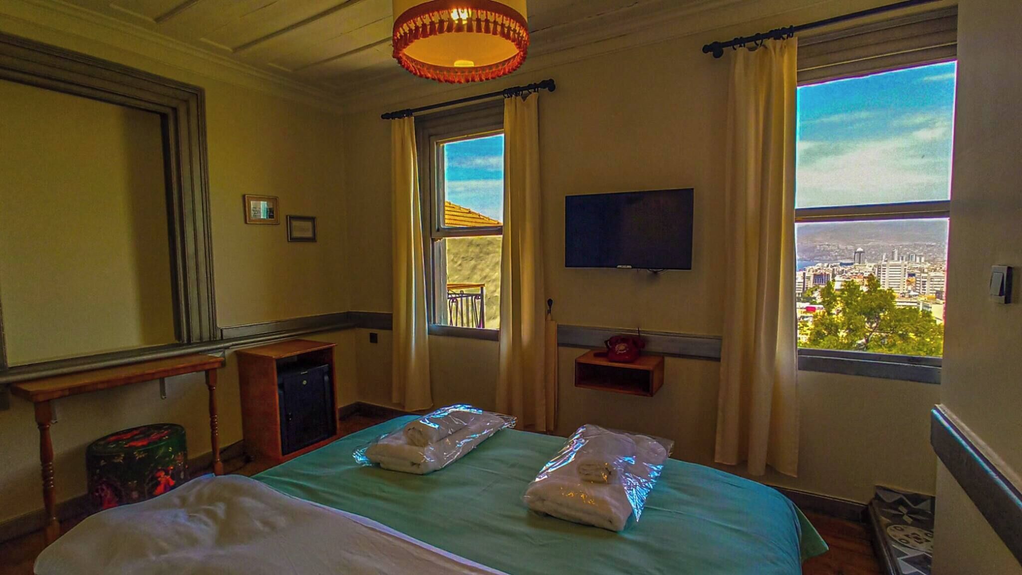 A hotel room with a bed, a television, a window with a city view, and a small table with a telephone.