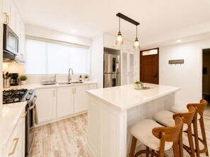 Cocina moderna con armarios blancos, una isla central con taburetes de bar, electrodomésticos de acero inoxidable e iluminación colgante.