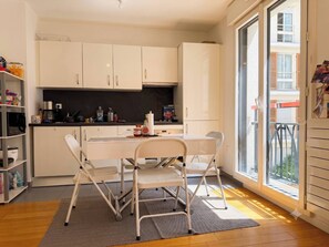 A modern kitchen with white cabinets, a dining table with chairs, and a view of the outside through a large window. - Charming Studio in the Heart of Paris - Ideal for Couples (Paris)