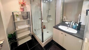 Un baño moderno con ducha envuelta en vidrio, tocador con lavamanos y repisa con artículos de aseo.