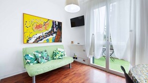 Een kamer met een groene bank, een flatscreen televisie, een schilderij en uitzicht op een groen gazon door een glazen schuifdeur.