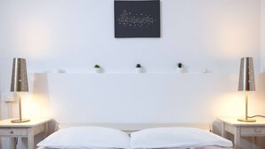 Een slaapkamer met een bed, twee nachtkastjes met lampen, een wandplank met kleine planten en een kunstwerk in een lijst boven het bed.