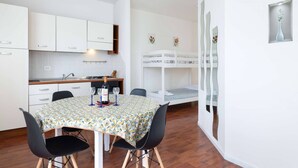 Een compacte keuken met witte kastjes, een eettafel met een tafelkleed met patroon, en een stapelbed met een spiegel.