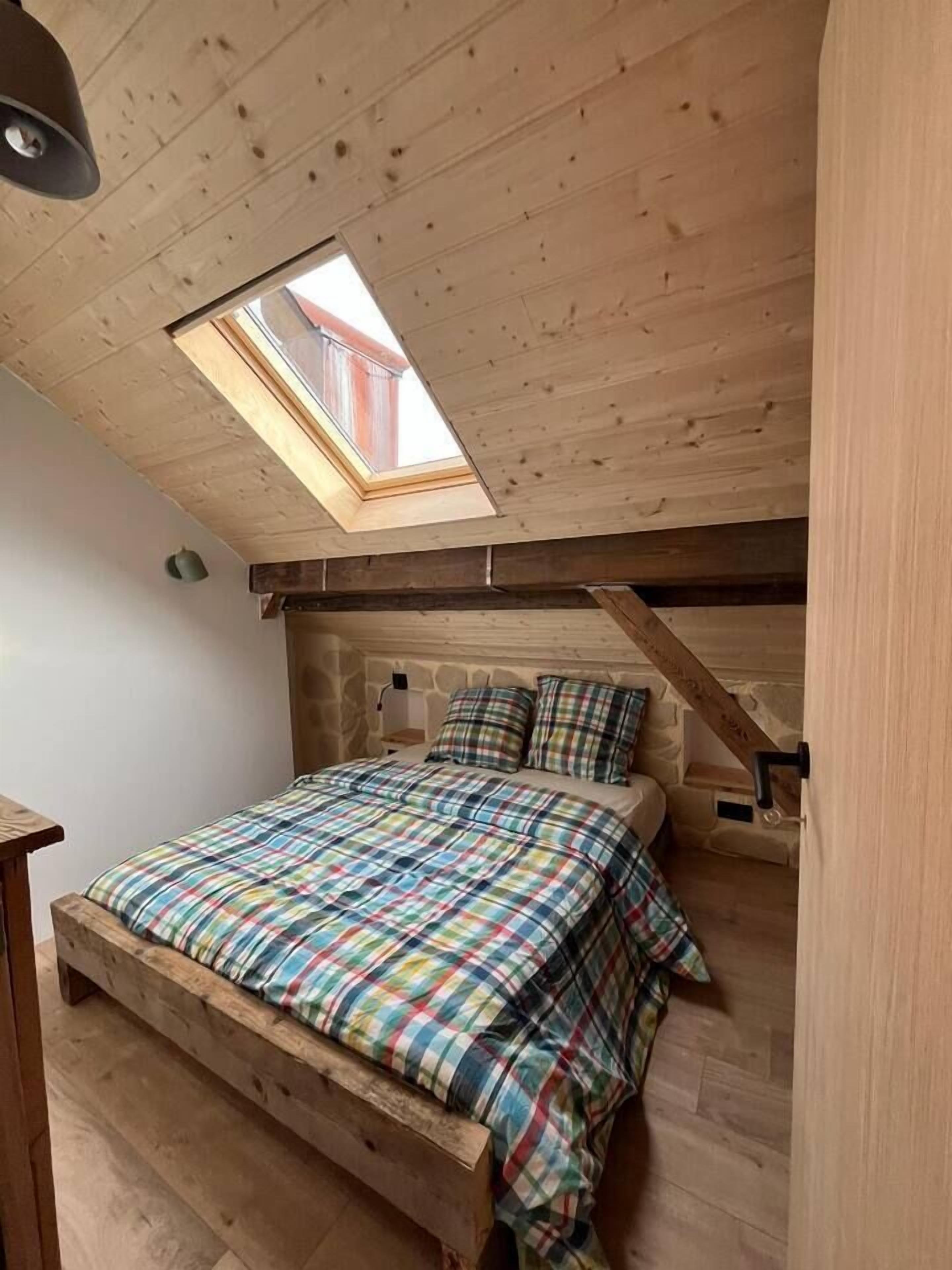 Ein Schlafzimmer mit schrägem Dach, einem Oberlicht, einem Bett mit kariertem Überwurf und einer Steinwand.