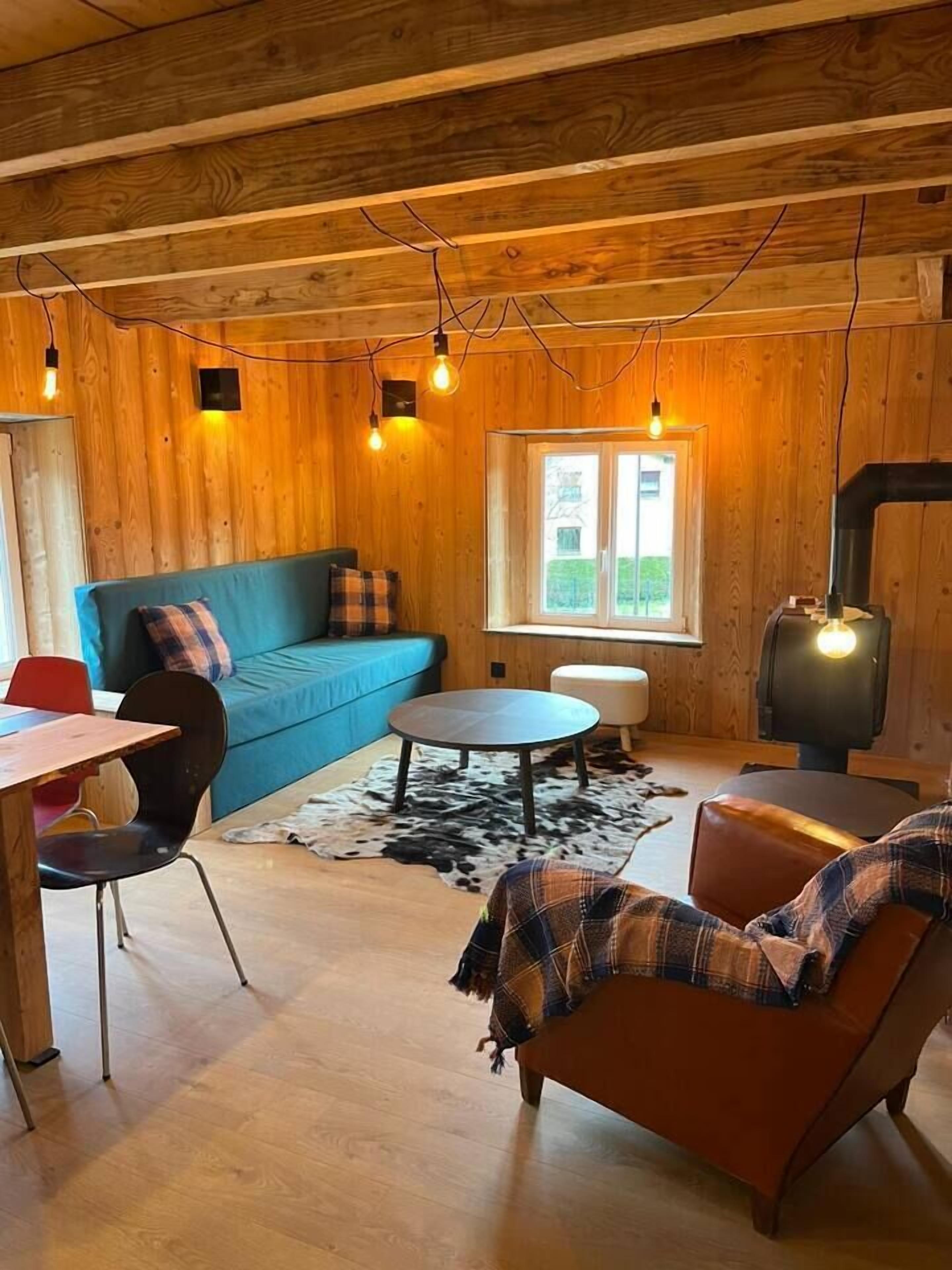 Ein gemütliches Blockhaus-Interieur mit Holzbalkendecke, einer blauen Couch, einem runden Tisch, einem Ledersessel und einem Kaminofen.