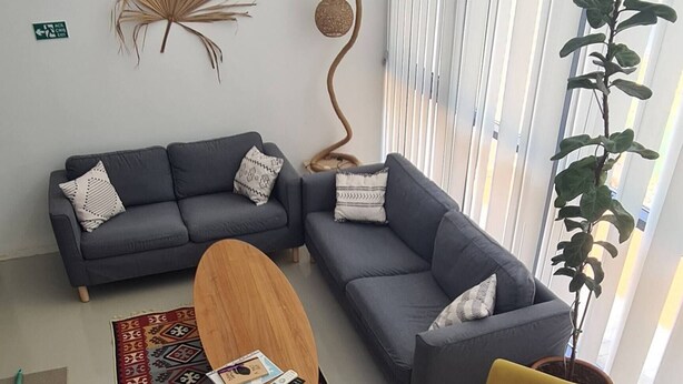 Uma sala de estar moderna com um sofá cinzento, uma poltrona amarela, uma mesa de centro de madeira e uma grande janela com persianas verticais.
