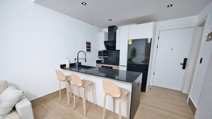 Une cuisine moderne avec un plan de travail noir, trois tabourets de bar en bois, un canapé blanc et une porte blanche.