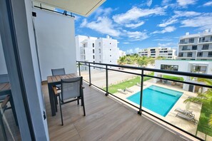 A balcony with a wooden table and chairs, overlooking a swimming pool and a landscaped area with palm trees. - Modern Apartment with Private Pool in Punta Cana (Punta Cana)