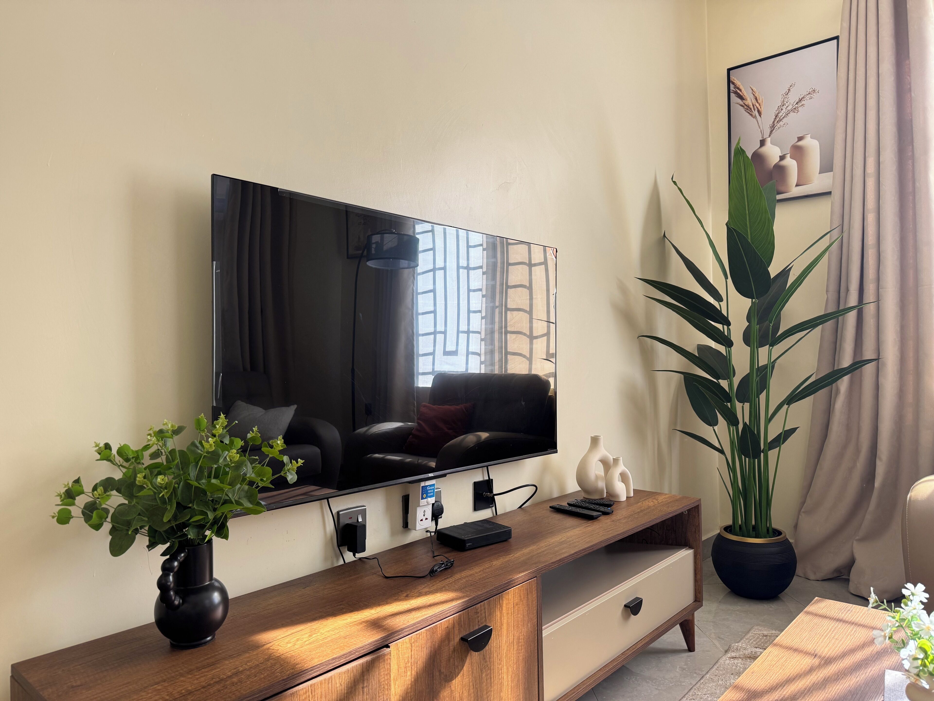 A modern living room with a flat-screen TV, a wooden console table, a potted plant, and a large window.