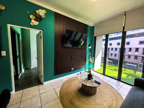 Ein modernes Wohnzimmer mit Flachbildfernseher an einer Wand aus Holzpaneelen, einem runden Couchtisch mit Fernbedienungen und Blick auf einen Gebäudekomplex durch große Fenster.
