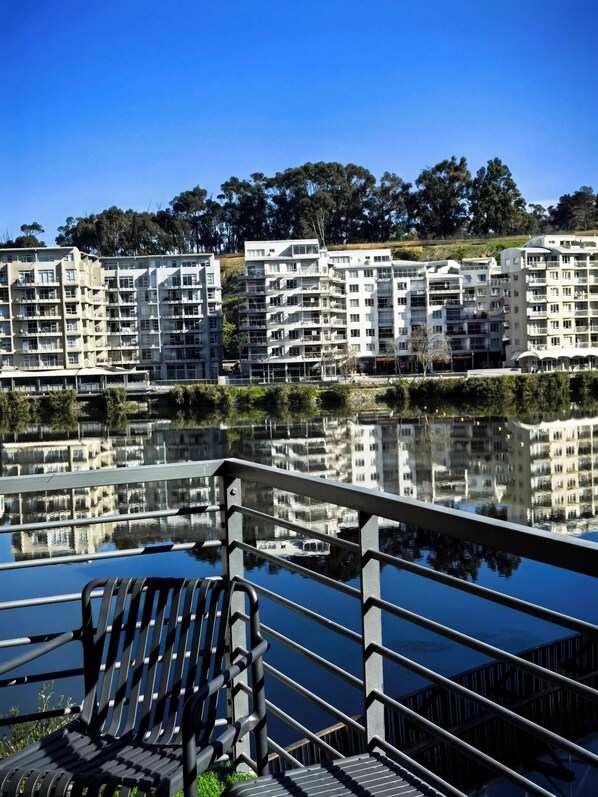 Ein Wohngebäude an der Uferpromenade mit Balkonen, ein Spiegelbild im Wasser und einem Metallgeländer im Vordergrund.