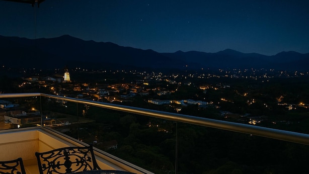 Un balcon avec une table et des chaises, une vue sur le paysage urbain illuminé la nuit et des montagnes à l’horizon.