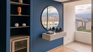 Ein Zimmer mit blauer Wand, einem runden Spiegel, einer Ablage mit Dekorationen, einem Schrank mit Glastüren und Blick auf Gebäude durch ein Fenster.