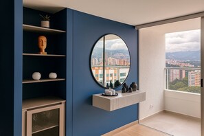 A room with a blue wall, a round mirror, a shelf with decorative items, a cabinet with glass doors, and a view of buildings through a window. - Epic View 25 • Pablo Tobón Cardiovascular • (Medellín)