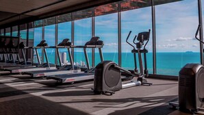 Et fitnesscenter med løbebånd, crosstrainere og en motionscykel, med udsigt til havet og bygninger gennem store vinduer.