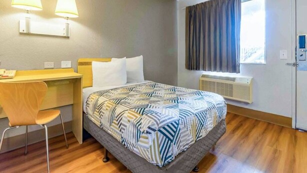 A hotel room with a bed, desk, chair, two wall-mounted lamps, a window with curtains, and an air conditioning unit. - 1 Queen Bed | Micfridge (Hayward)