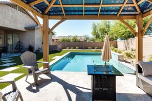 A covered outdoor swimming pool area with a lounge chair, a table with an umbrella, and a patio table. - 5 Mi to Estrella Mtn Park! Home w/ Outdoor Oasis (Goodyear)