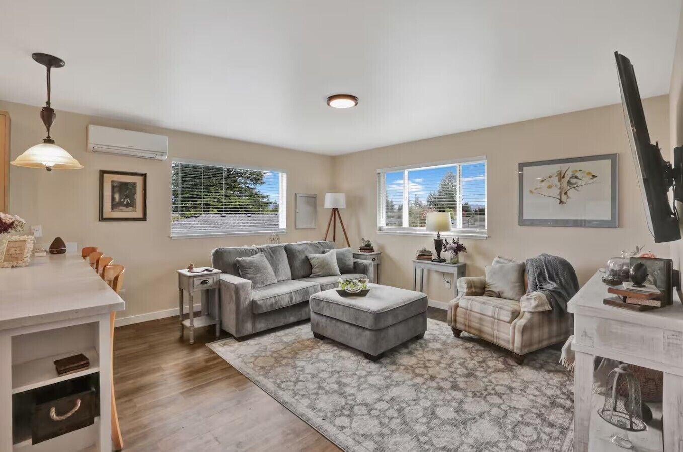A spacious living room with a grey sofa, a matching ottoman, and a plaid armchair. There is a dining area with a table and chairs, and a large window providing a view of the outdoors.