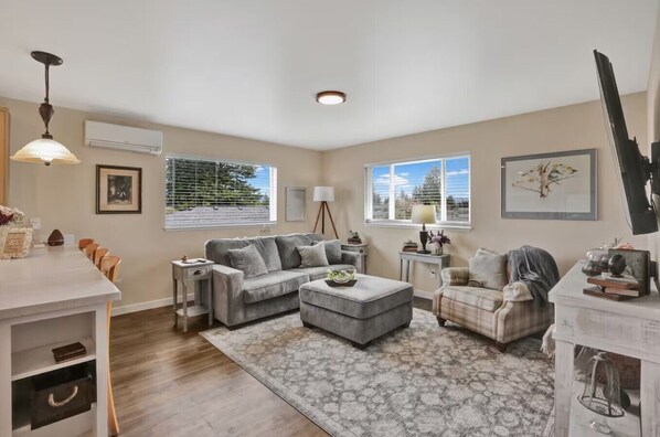 A spacious living room with a grey sofa, a matching ottoman, and a plaid armchair. There is a dining area with a table and chairs, and a large window providing a view of the outdoors.