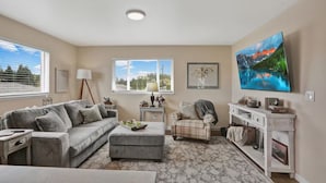 A living room with a grey sofa, a small table, a plaid armchair, a flat-screen TV, and a large window with blinds. - The Nest House (Burlington)