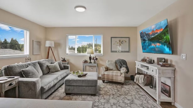 A living room with a grey sofa, a small table, a plaid armchair, a flat-screen TV, and a large window with blinds. - The Nest House (Burlington)
