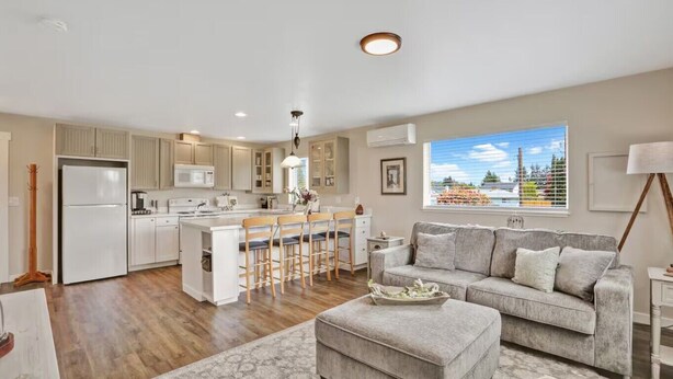 A modern kitchen with a central island, bar stools, a grey sofa, and a view of a cityscape through a large window. - The Nest House (Burlington)