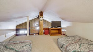 Une chambre mansardée avec un plafond en bois, une horloge, un téléviseur posé sur un meuble en bois et deux lits avec une literie à motifs.
