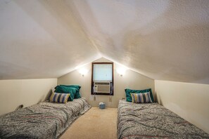 Une petite chambre mansardée de forme triangulaire, avec deux lits simples, une fenêtre avec des stores et un climatiseur fixé au mur.