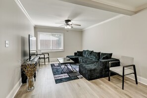 Ein Wohnzimmer mit Ecksofa, Couchtisch, Sessel, einer Vase mit Trockenblumen, Deckenventilator und einem Fenster mit Jalousien.