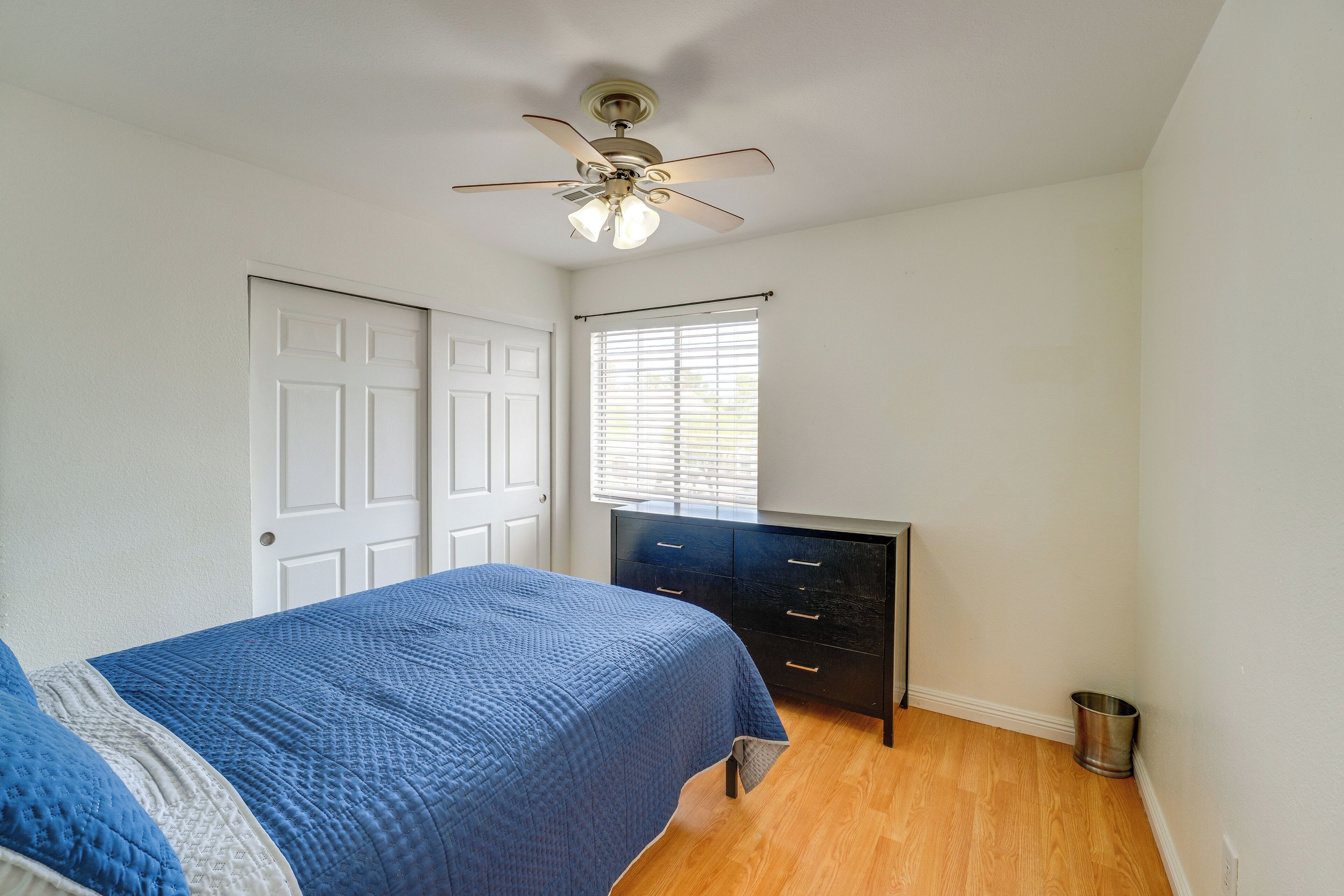 A bedroom with a bed, a dresser, a ceiling fan, a window with blinds, and a door.