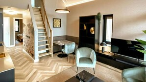 A modern living room with a herringbone floor, a staircase, a flat-screen TV, and a seating area with a light blue armchair.