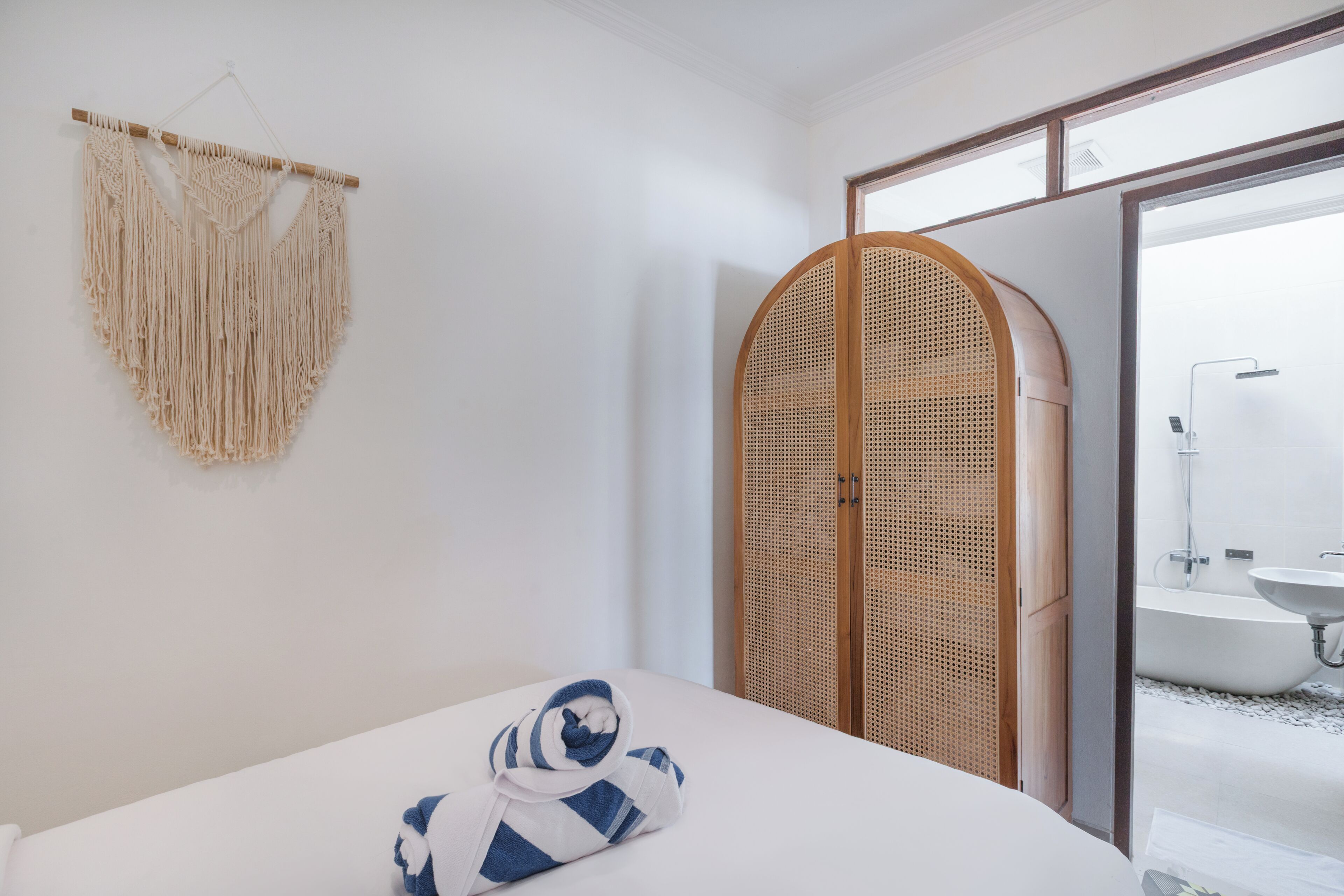 A bedroom with a bed, a macrame wall hanging, a rattan wardrobe, and a bathroom with a sink and shower.