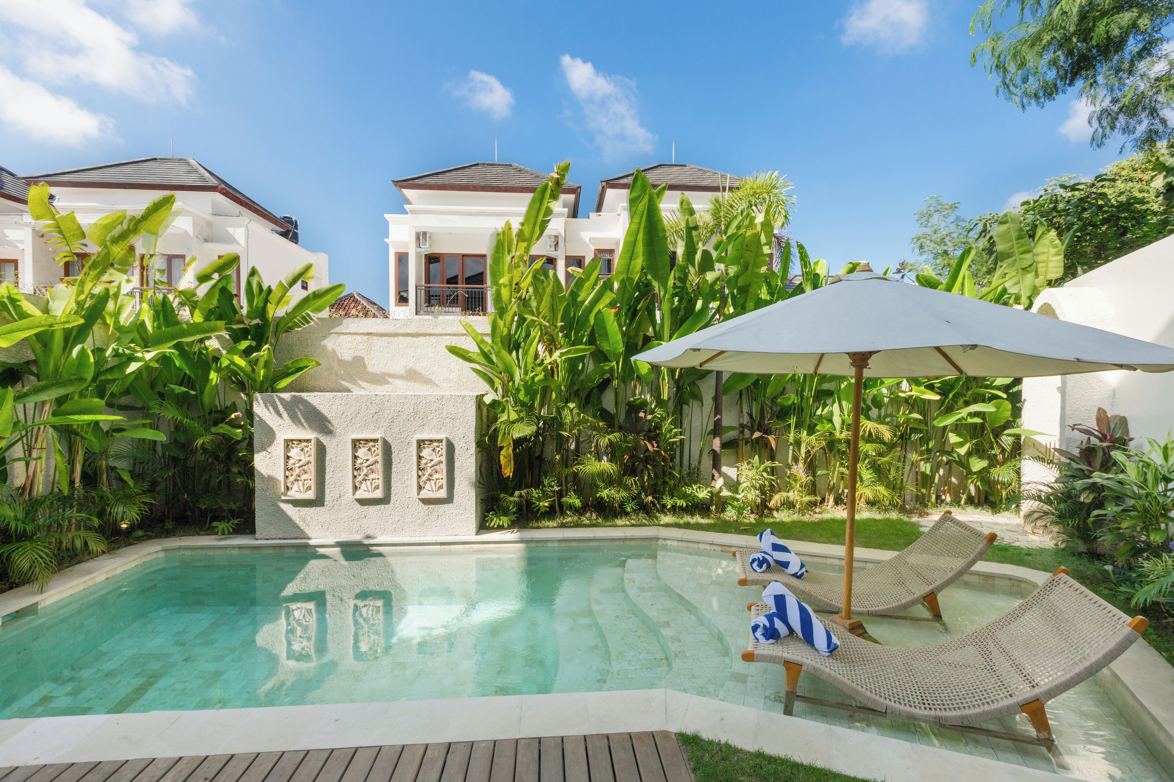 A swimming pool with lounge chairs and an umbrella, surrounded by tropical plants and a white building with a sloped roof.