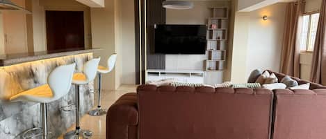 A modern living room with a sectional sofa, a bar area with stools, a flat-screen TV, and a built-in shelf unit.