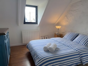 Ein Schlafzimmer mit schrägverdeckter Decke, einem Fenster, einem Heizkörper, einem Nachttisch mit Lampe, einem Bett mit gestreifter Bettwäsche und einem blauen Kleiderschrank.