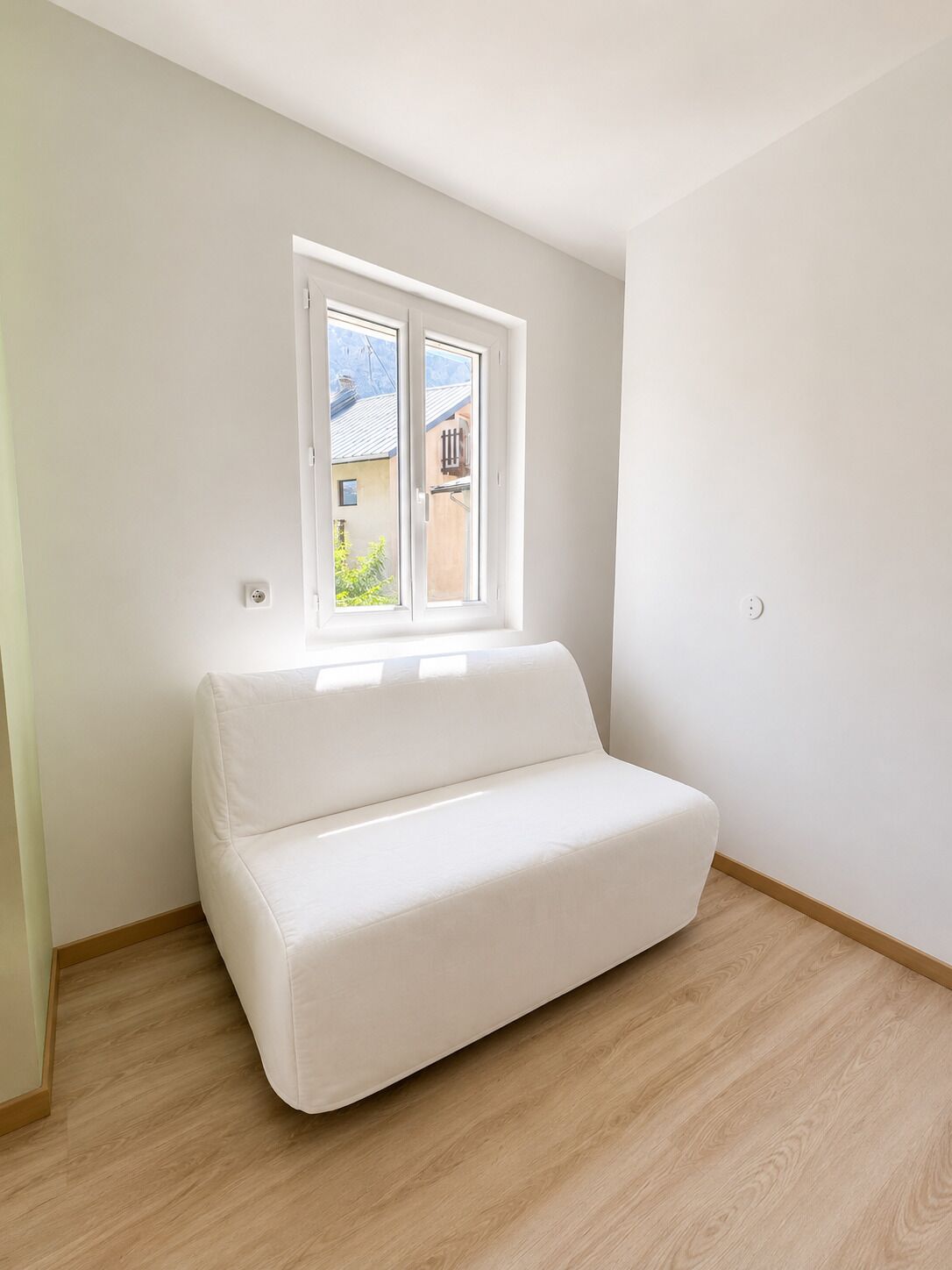 Un petit canapé blanc, à une place, se trouve dans une pièce dont le sol est en bois et qui possède une fenêtre donnant sur des bâtiments à l’extérieur.