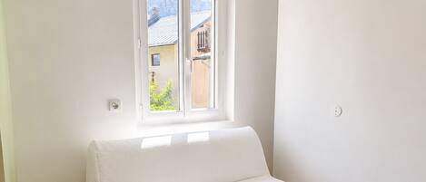Un petit canapé blanc, à une place, se trouve dans une pièce dont le sol est en bois et qui possède une fenêtre donnant sur des bâtiments à l’extérieur.
