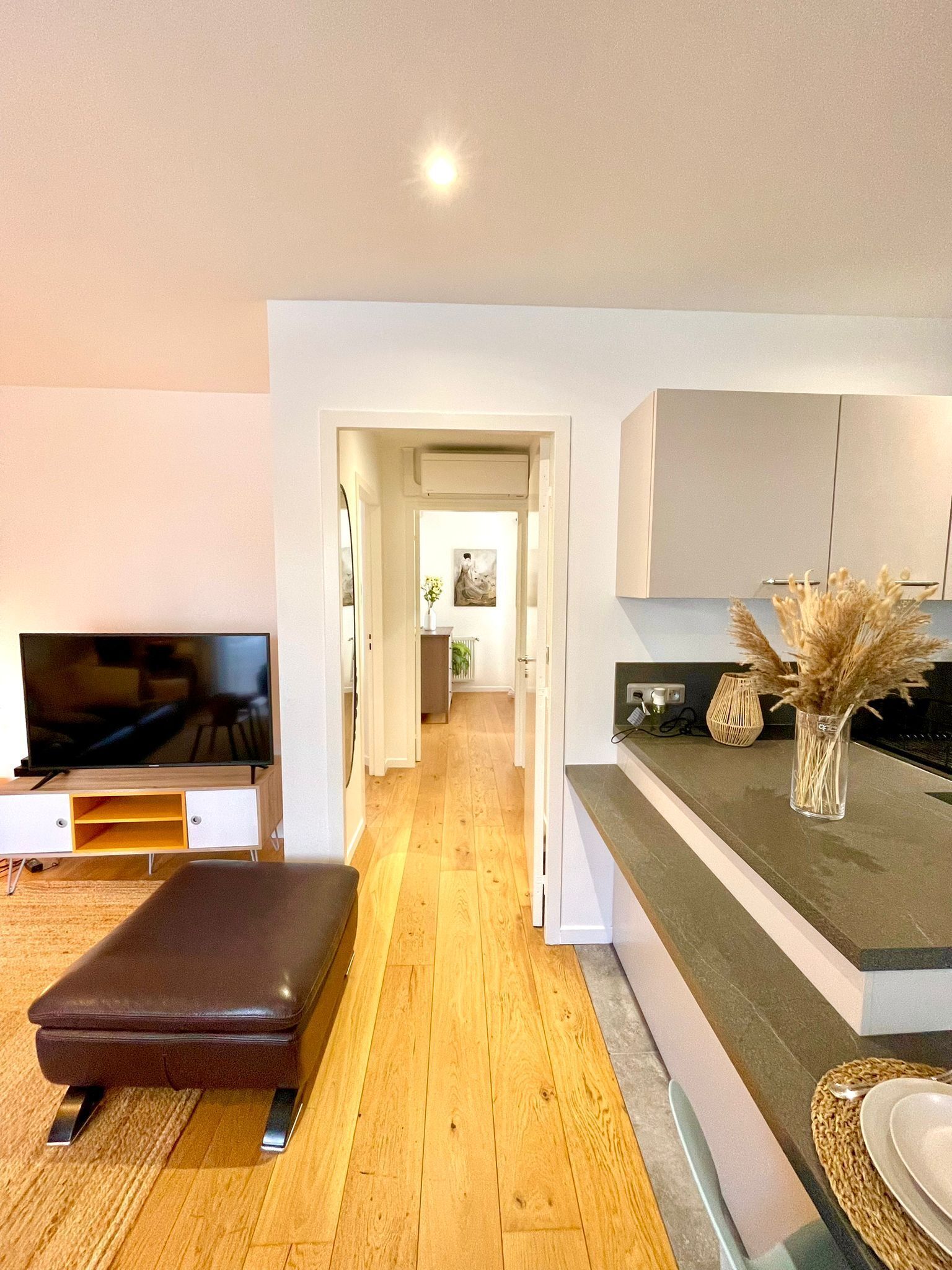 Una sala de estar moderna con un televisor de pantalla plana, un reposapiés de cuero y una zona de cocina con un jarrón de plantas secas.