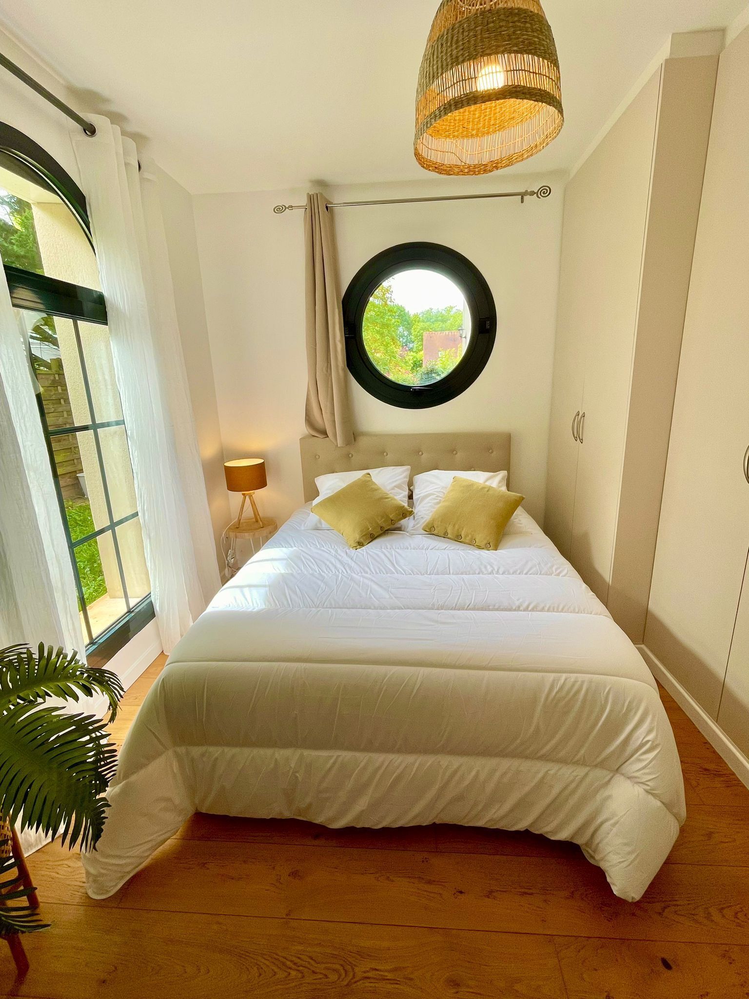 Un dormitorio con cama, un ventanal redondo, una lámpara colgante y una planta.