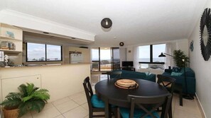 A modern living room with a dining table, chairs, and a sofa. There is a kitchen area with a window and a view of the ocean. - Palm Beach Stunning Ocean Apartment! (Palm Beach)