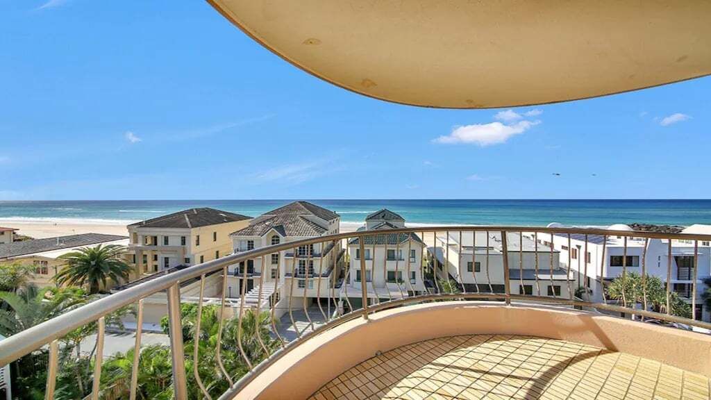 A balcony with a view of the beach and ocean, a curved railing, and a building with multiple floors and balconies.