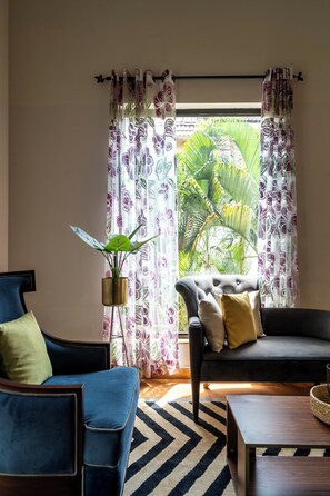A living room with a sofa, a coffee table, a potted plant, and a view of tropical foliage through patterned curtains. - Staymaster Trivala-3BR Villa with Pool  (Calangute)