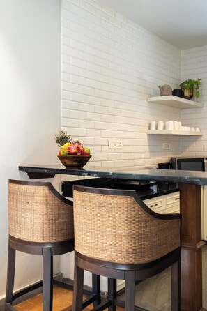 A kitchen with white brick walls, a black countertop, wicker bar stools, and a bowl of fruit. - Staymaster Trivala-3BR Villa with Pool  (Calangute)