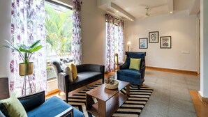 Un salon comprenant un canapé, un fauteuil et une table basse, ainsi qu’une plante en pot et des rideaux à motifs floraux.