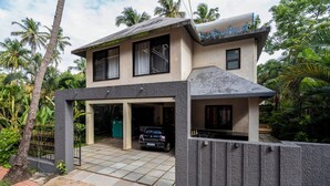 Une maison à deux étages avec une voiture garée dans l’allée, entourée d’une végétation luxuriante et de palmiers.