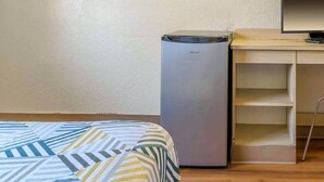 A room with a bed, a small refrigerator, a desk with a computer, and a shelf unit. - 1 Queen Bed | Hayward (Hayward)
